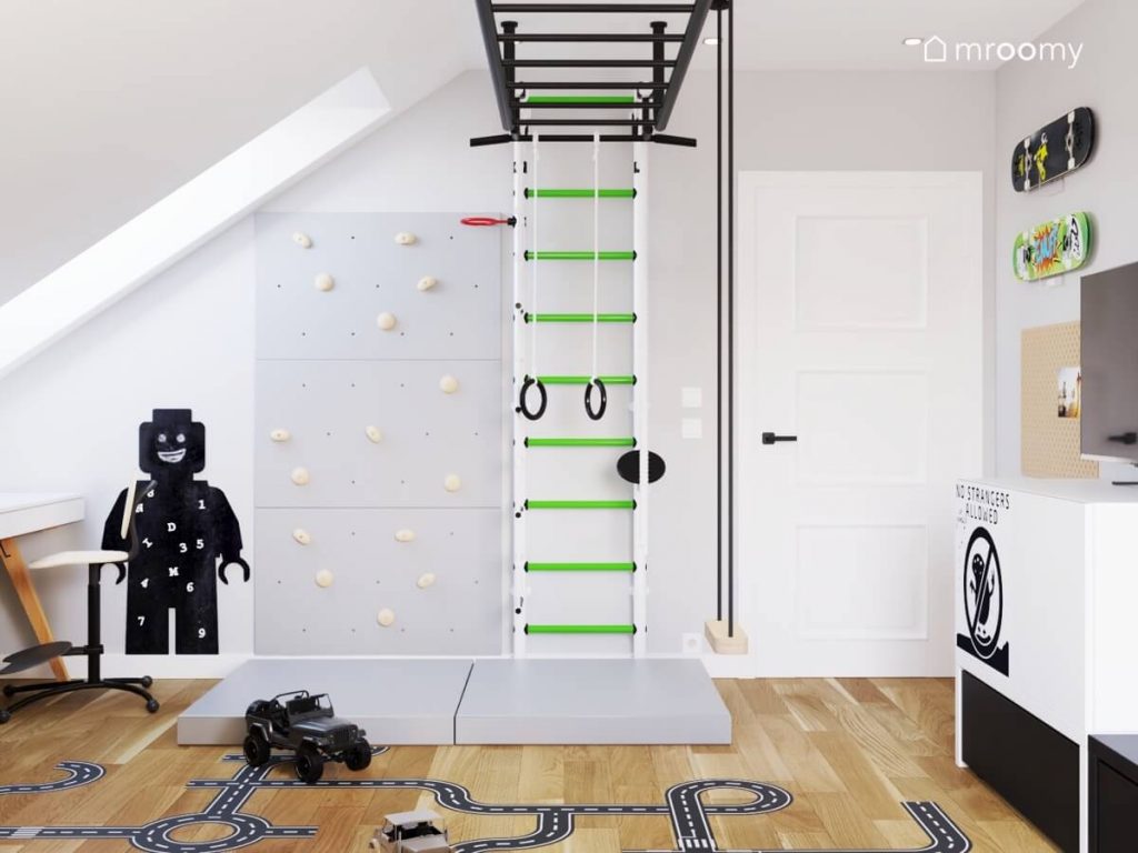 Czarna tablica kredowa w kształcie ludzika Lego szara ścianka wspinaczkowa biało zielona drabinka gimnastyczna a na suficie czarna drabinka sufitowa oraz huśtawka w pokoju chłopca w wieku szkolnym