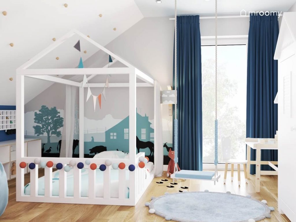 Duże białe łóżko domek uzupełnione girlandą cotton balls oraz proporczykami a za nim tapeta z motywem gospodarstwa a na środku niebieska huśtawka wisząca w pokoju małego chłopca