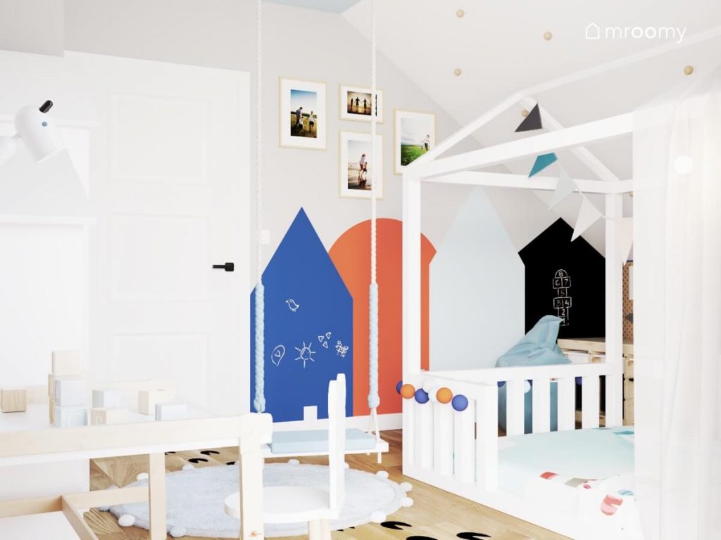 Białe łóżko domek i inne białe meble a u sufitu zawieszona błękitna huśtawka a na ścianie powierzchnie kredowe w kształcie domków i zdjęcia w poddaszowym pokoju chłopca