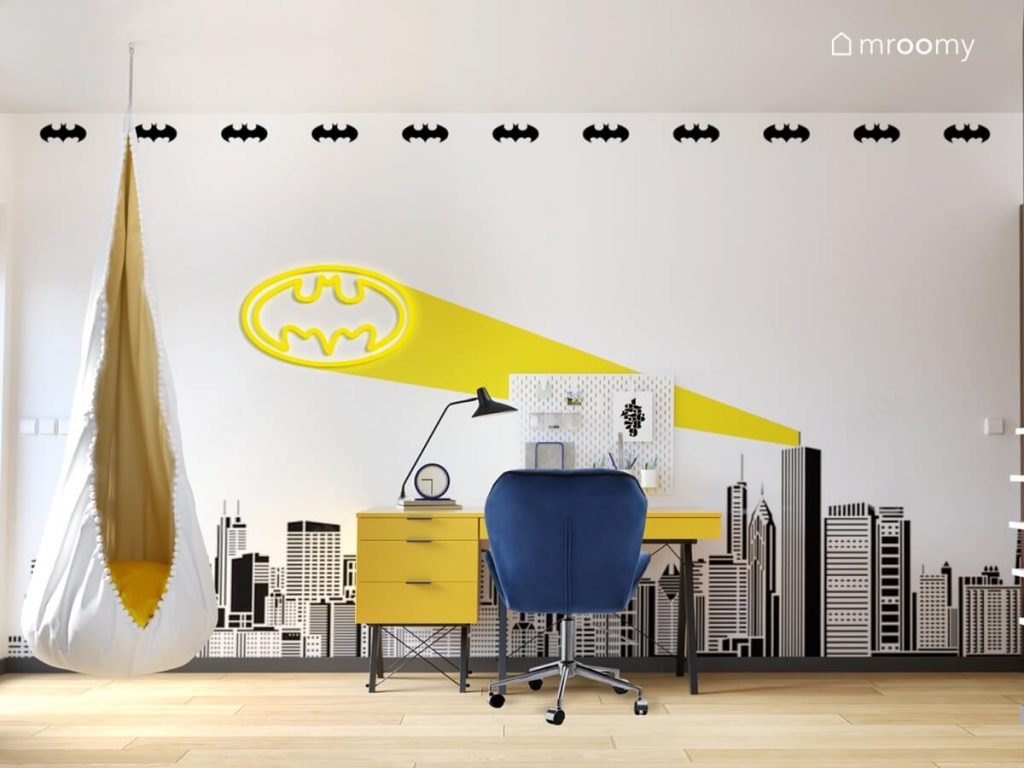 Żółte biurko z kontenerkiem i niebieskim krzesłem a za nią ściana pokryta tapetą z panoramą miasta i symbolami Batmana a na niej ledon z Bat symbolem a obok biało żółty fotel kokon w pokoju chłopca