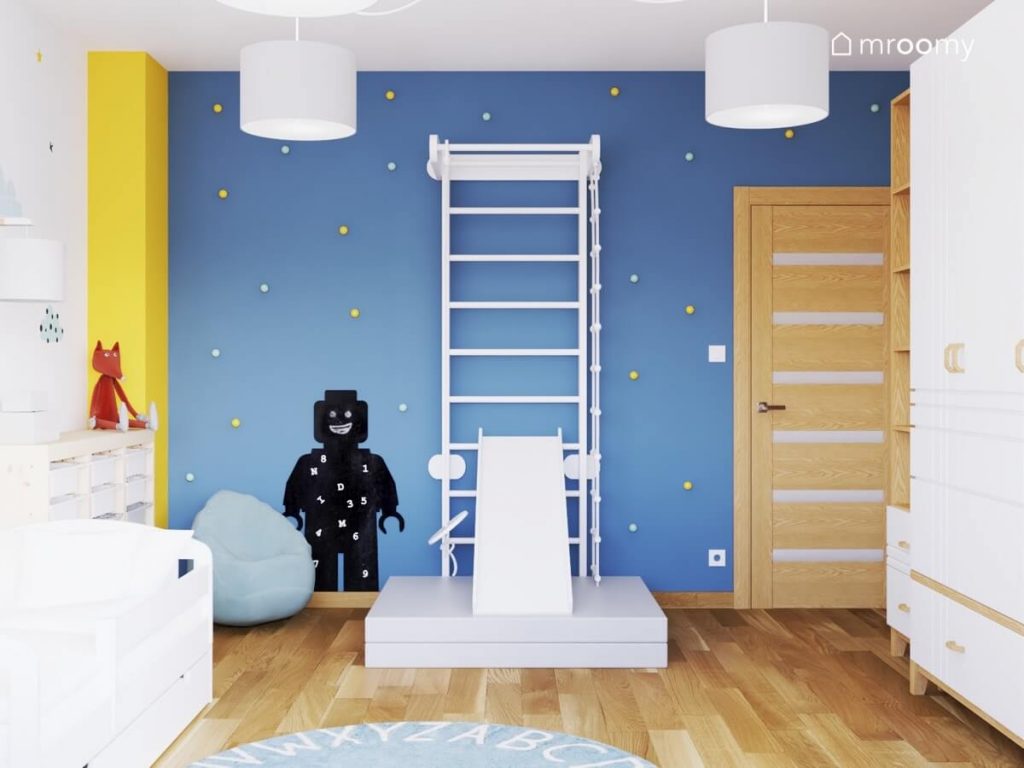 Niebieska ściana w pokoju chłopca w wieku przedszkolnym uzupełniona żółtymi i błękitnymi gałkami a na niej biała drabinka gimnastyczna i tablica kredowa w kształcie ludzika Lego