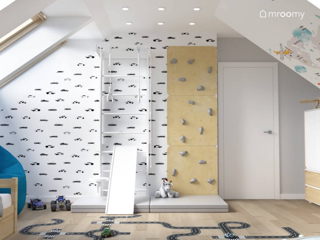 Ściana w pokoju dla chłopca pokryta tapetą w samochody a na niej biała drabinka gimnastyczna i drewniana ścianka wspinaczkowa