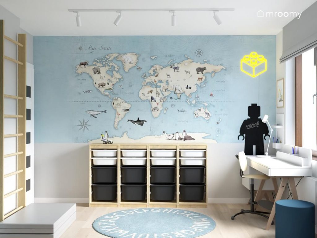 Ściana w pokoju chłopca pokryta tapetą z mapą świata a na niej drewniany regał z pojemnikami tablica kredowa w kształcie ludzika Lego oraz ledon klocek
