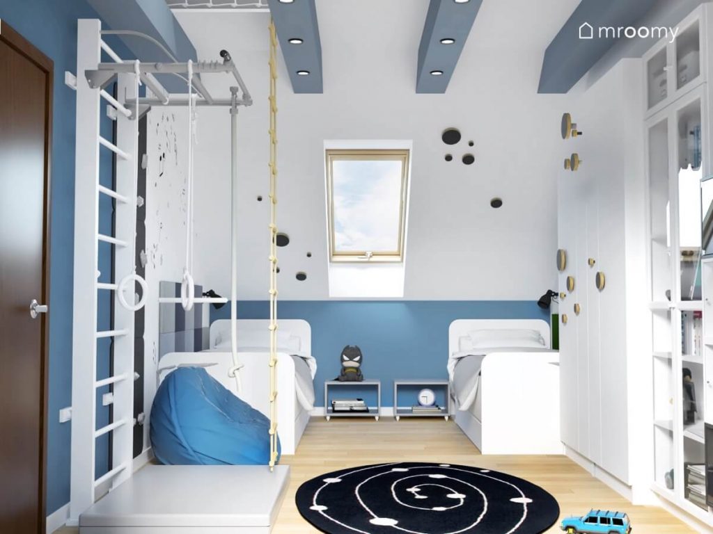 Biało niebieski pokój dla dwóch chłopców a w nim dwa białe łóżka drabinka gimnastyczna niebieska pufa oraz skos i szafa uzupełnione wieszakami ze sklejki