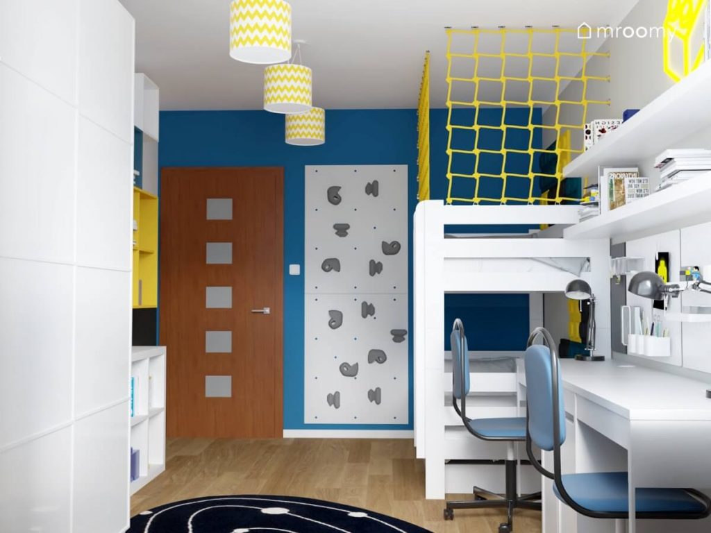 Biało szaro niebieski pokój dwóch chłopców a w nim ścianka wspinaczkowa łóżko piętrowe oraz dwa białe biurka z niebieskimi krzesłami