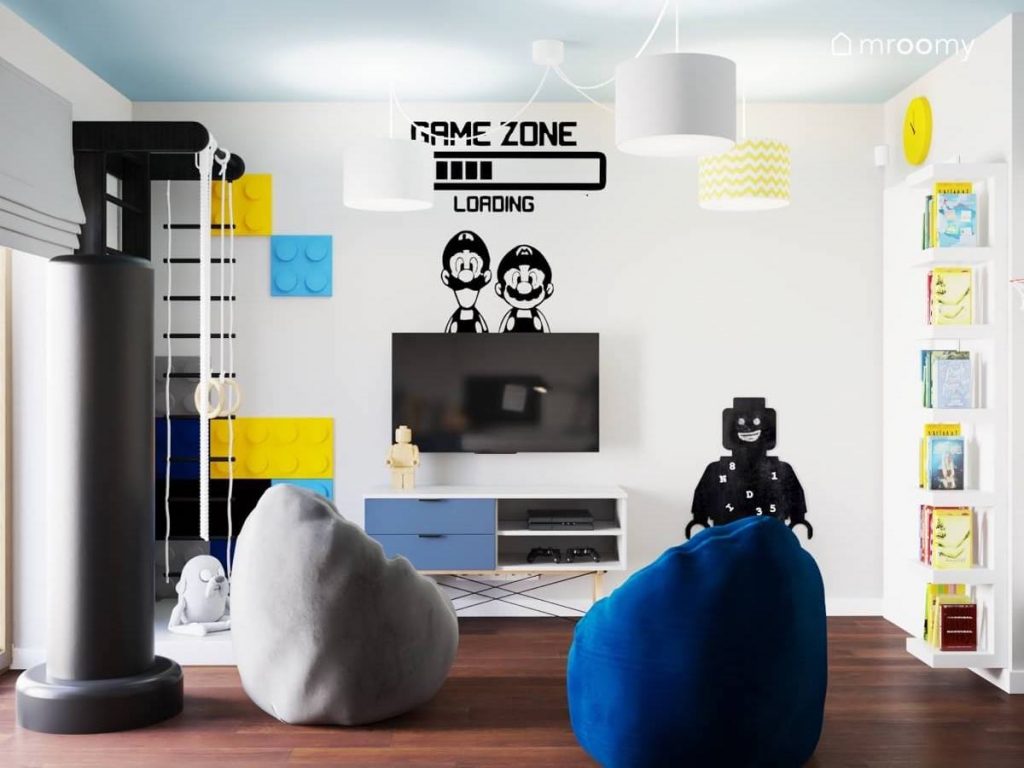 Strefa rozrywki w pokoju dwóch chłopców a w niej telewizor szafka rtv szara i niebieska pufa a także gamingowa naklejka i tablica kredowa w kształcie ludzika Lego