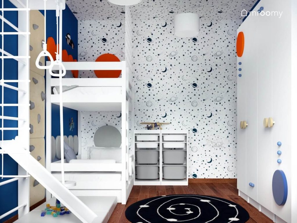 Ściana i sufit oklejone biało niebieską kosmiczna tapetą a także białe łóżko piętrowe i regał z pojemnikami na zabawki a na podłodze galaktyczny dywan w pokoju dwóch braci