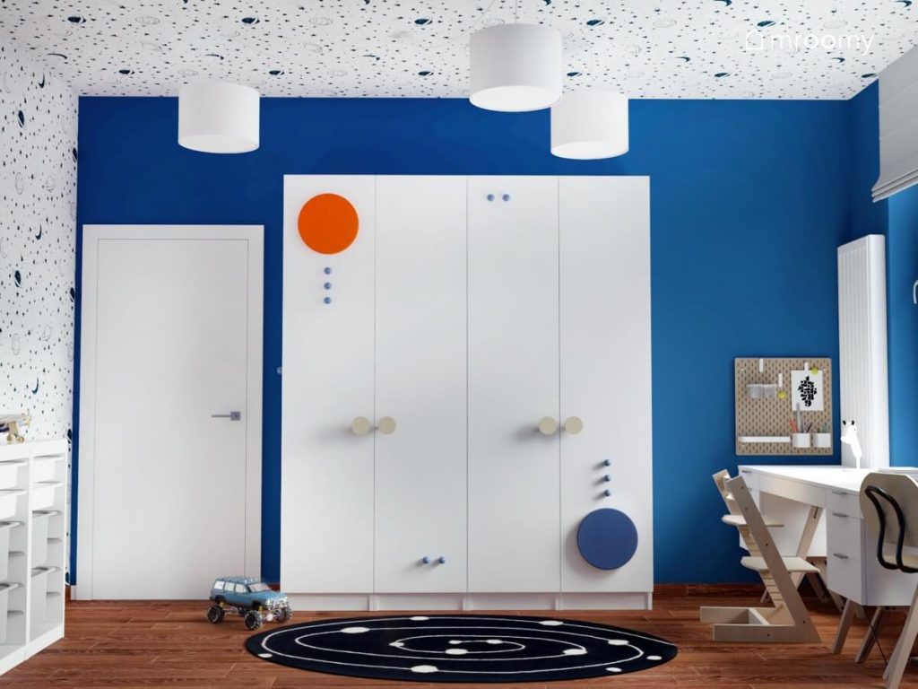Duża biała szafa uzupełniona pomarańczowym i niebieskim panelem oraz drewnianymi i niebieskimi gałkami a u sufitu białe lampy w niebieskim pokoju dla chłopców