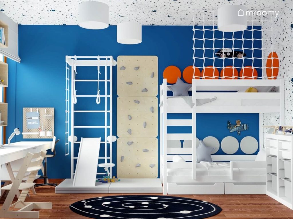Biała drabinka gimnastyczna ze zjeżdżalnią ścianka wspinaczkowa i białe łóżko piętrowe w intensywnie niebieskim pokoju dwóch chłopców