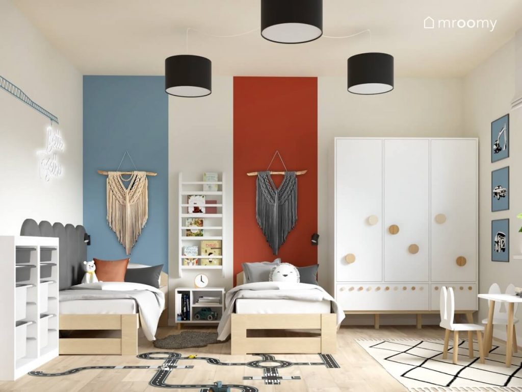 Dwa drewniane łóżka a na ścianie ozdobne makramy i pasy farby w kolorze niebieskim i rdzawym a między nimi biblioteczka i szafka nocna a obok biała szafa z wieszakami ze sklejki w pokoju dla dwóch chłopców