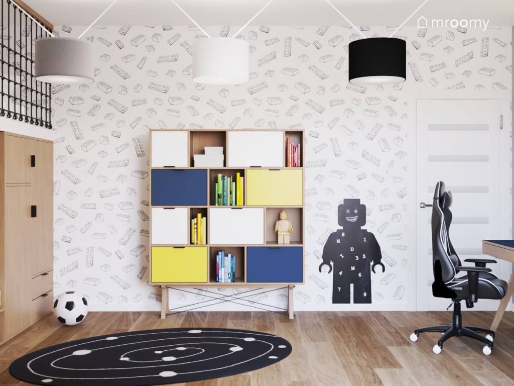 Drewniany regał w kolorowymi szufladami a także tablica kredowa w kształcie ludzika Lego galaktyczny dywan i tapeta w klocki w pokoju dwóch chłopców