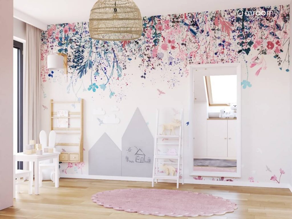 Ściana pokryta tapetą w kwiaty a na niej drewniana biblioteczka tablice kredowe w kształcie domków prostokątne lustro oraz regał drabinka w pokoju dwóch dziewczynek
