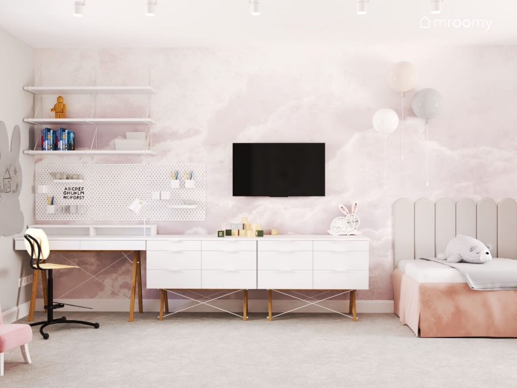 Ściana pokryta wielkoformatową fototapetą z motywem chmur a na niej białe półki i telewizor a oprócz tego białe meble na drewnianych nogach w pokoju dziewczynki