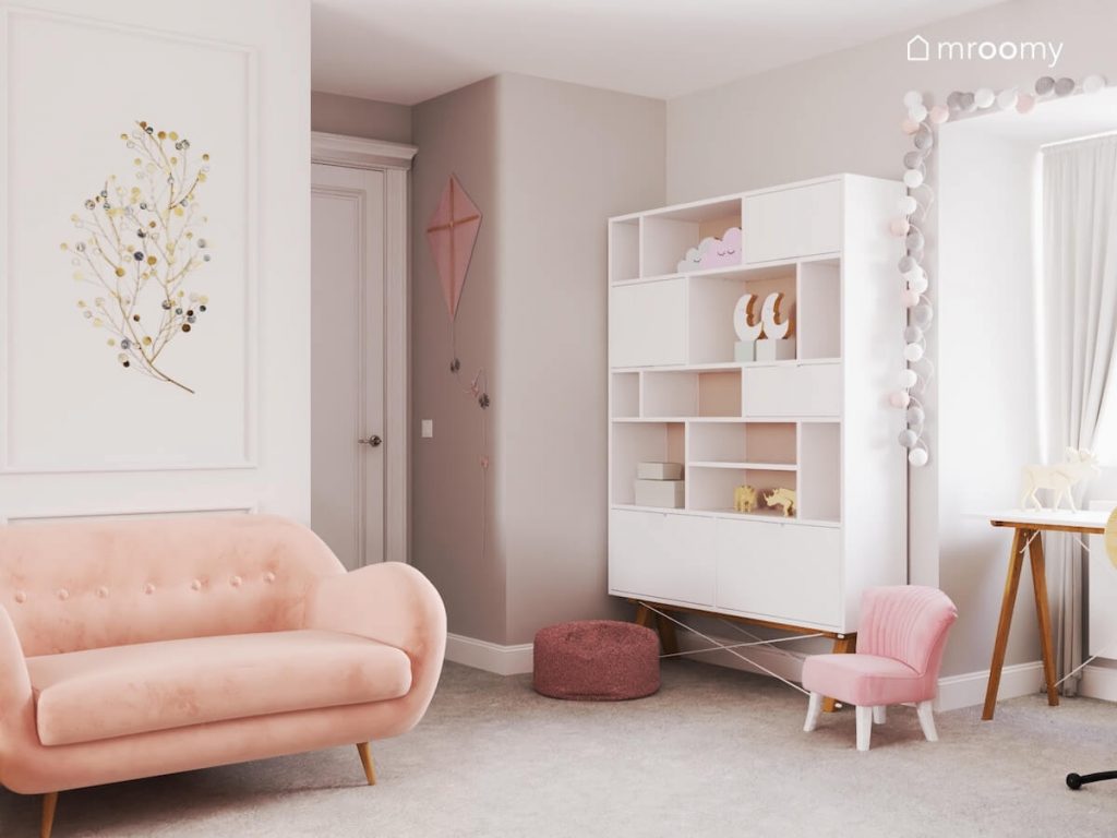 Różowa kanapa biały regał na drewnianych nogach różowy fotelik i pufa a ściany ozdobione girlandą cotton balls oraz dekoracjami w kształcie latawca i gałązek w pokoju dziewczynki