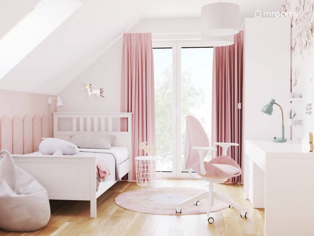 Białe łóżko a nad nim lampka w kształcie jednorożca a także białe biurko z różowym krzesłem i różowe zasłony w poddaszowym pokoju dziewczynki