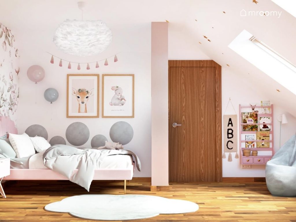 Różowe tapicerowane łóżko z ozdobnym zagłówkiem uzupełnione okrągłymi szarymi panelami girlandą pomponów oraz ozdobnymi plakatami ze zwierzętami a także różowa biblioteczka i niebieska pufa w pokoju dla dziewczynki