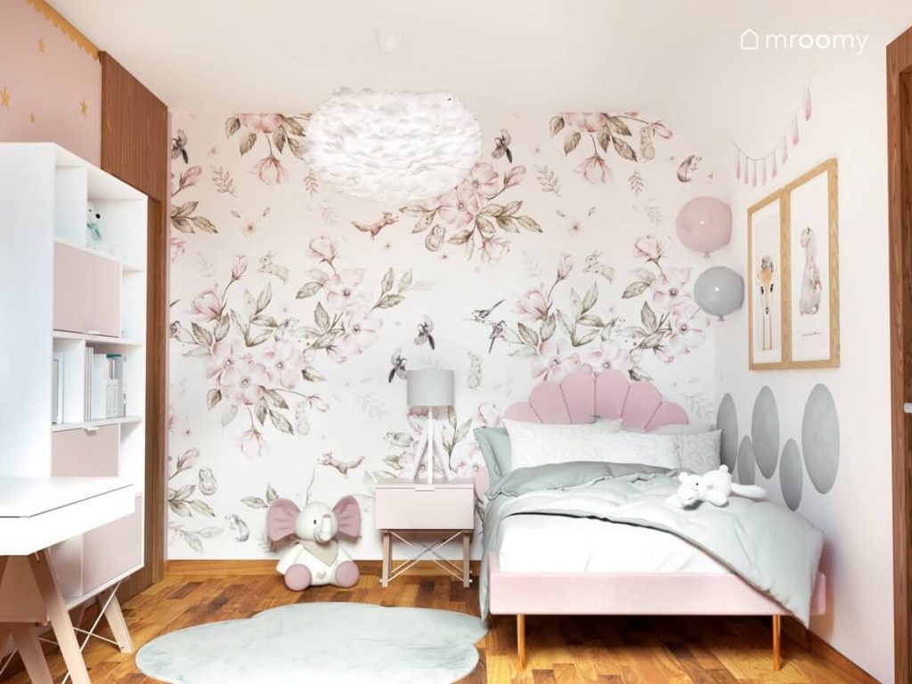 Jasny przytulny pokój dla dziewczynki w wieku szkolnym a w nim różowe tapicerowane łóżko z ozdobnym zagłówkiem kinkiety w kształcie balonów lampa sufitowa z ptasich piór i piękna tapeta w kwiaty