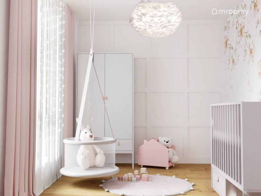 Biały pokój dla dziewczynki niemowlaka a w nim jasne meble różowa biblioteczka w kształcie domku huśtawka kojec oraz lampa z ptasich piór