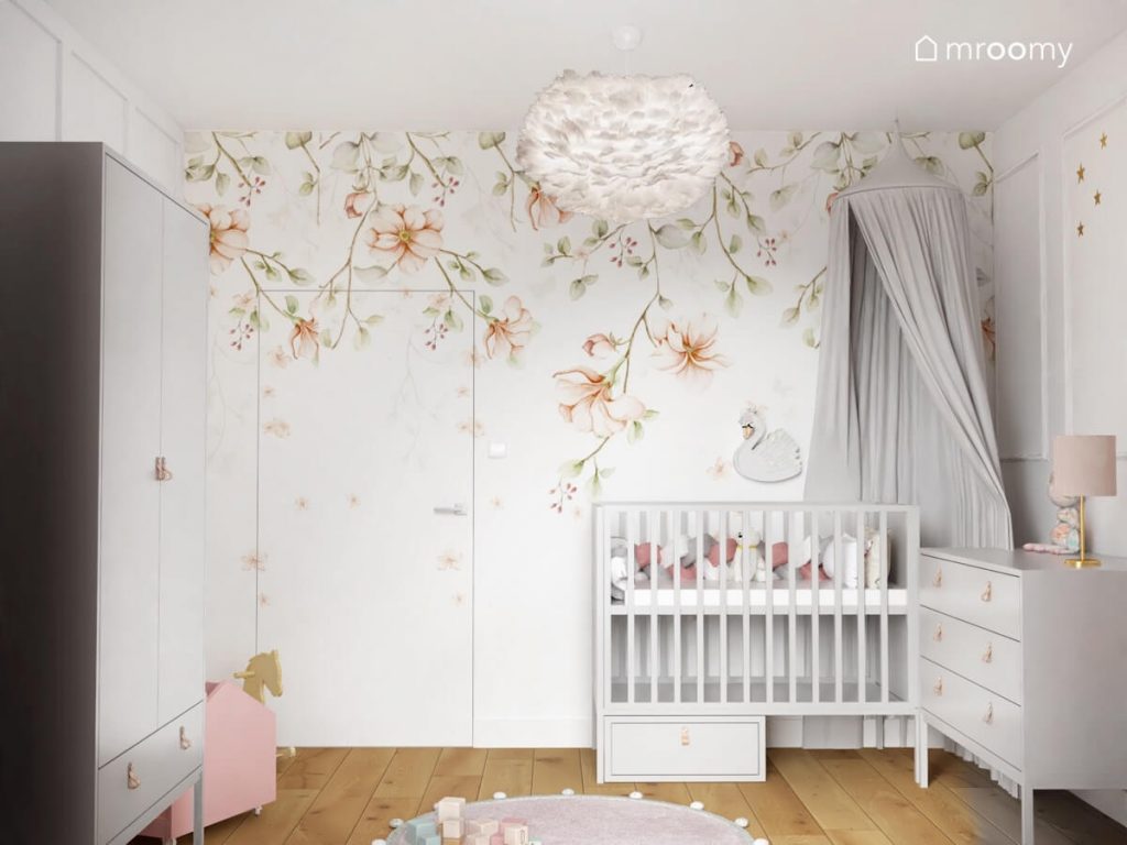 Ściana oklejona piękną tapetą w kwiaty a także łóżeczko dla niemowląt udekorowane szarym baldachimem a nad nim lampka nocna w kształcie łabędzie w pokoju dla malutkiej dziewczynki