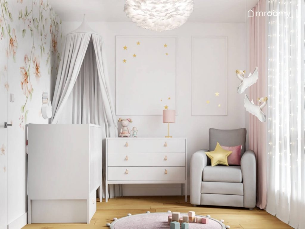 Łóżeczko dziecięce uzupełnione szarym baldachimem biała komoda oraz szary fotel a na ścianie ozdobna sztukateria uzupełniona złotymi gwiazdkami w pokoju dla niemowlaka