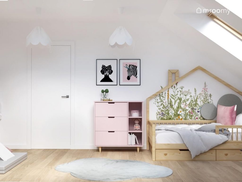 Drewniane łóżko w kształcie domku uzupełnione naklejką z kwiatami a także różowa komoda a nad nią plakaty ze zwierzętami a na suficie lampy bezy w poddaszowym pokoju sześciolatki