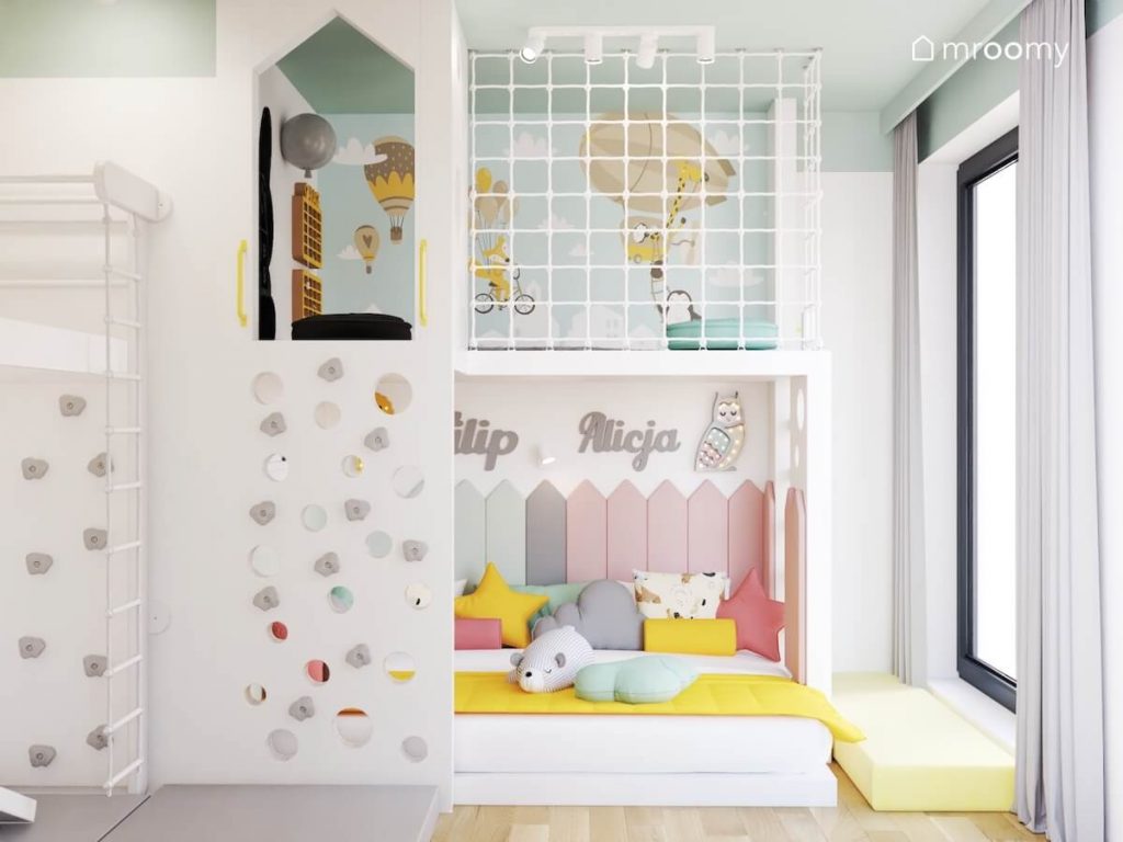 Biała antresola w pokoju dziewczynki i chłopca na którą prowadzi ścianka wspinaczkowa a na dole znajduje się miejsce na odpoczynek wyłożone miękkimi panelami ściennymi w kształcie płotka
