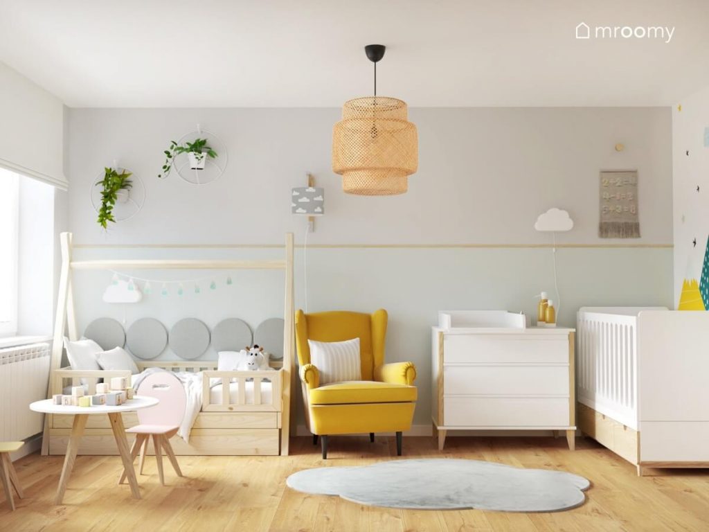 Biało szary pokój dla chłopca i dziewczynki z łóżeczkiem dla małego dziecka drewnianym łóżkiem tipi uzupełnionym panelami ściennymi a także żółtym fotelem i przewijakiem