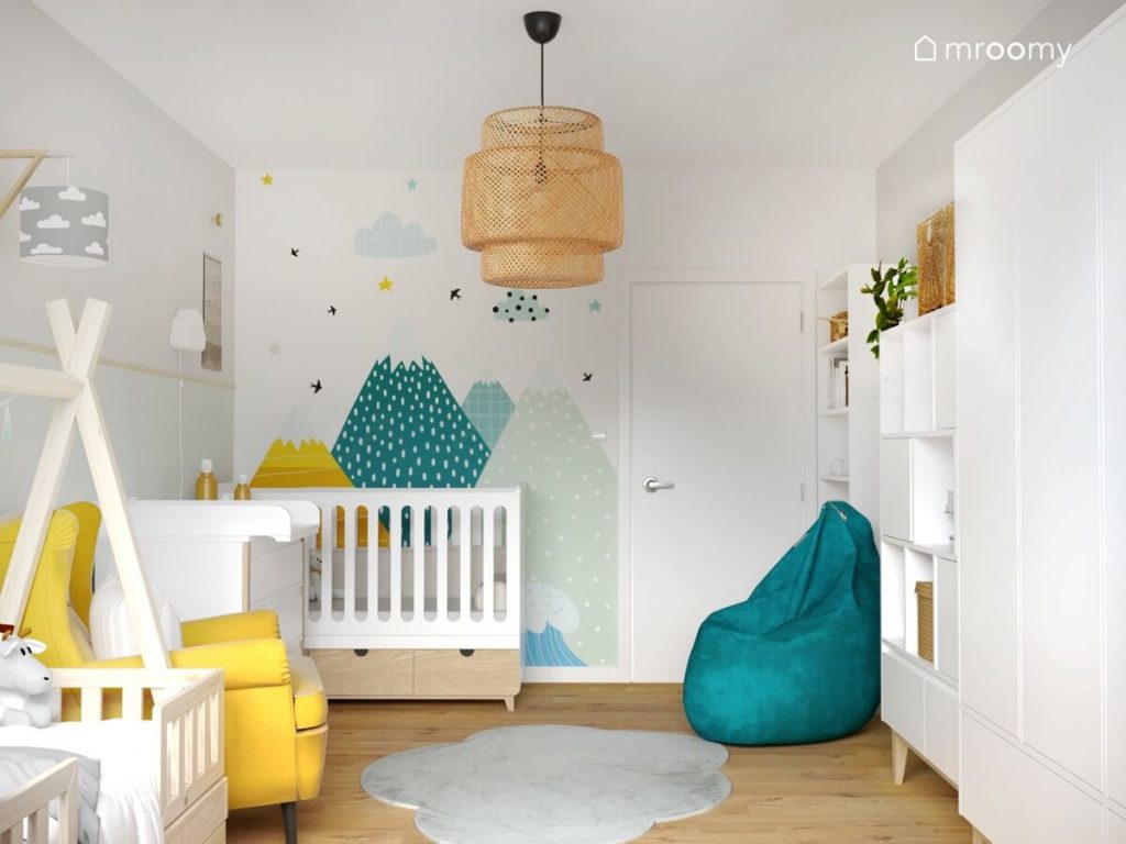 Jasny pokój dla dwójki małych dzieci z białymi i drewnianymi meblami turkusową pufą żółtym fotelem oraz tapetą z górzystym motywem