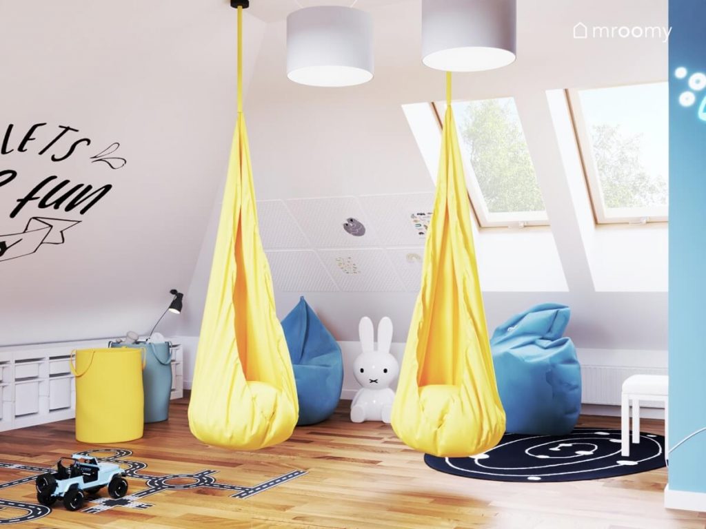 Dwa żółte wiszące fotele dwie niebieskie pufy a także worki na zabawki w tych samych kolorach a na podłodze galaktyczny dywan i lampa w kształcie królika w pokoju trójki rodzeństwa