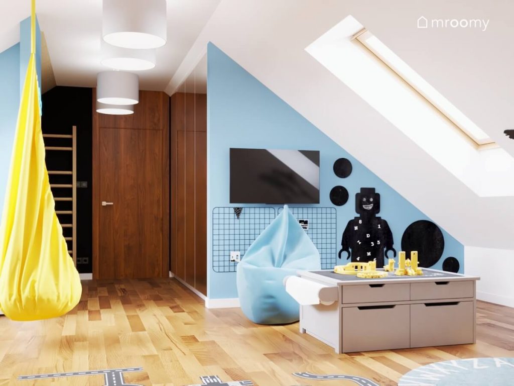 Strefa rozrywki w biało niebieskim poddaszowym pokoju rodzeństwa a w niej telewizor niebieska pufa stolik do zabawy oraz tablice kredowe na ścianie