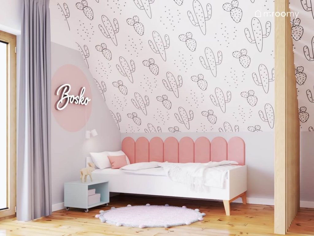 Różowa strefa spania dziewczynki w pokoju dla siostry i brata a w niej białe łóżko uzupełnione różowymi panelami ściennymi błękitna szafka nocna dywanik ledon w kształcie napisu a na skosie tapeta w kaktusy
