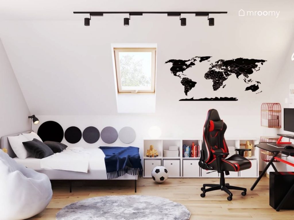 Jasny poddaszowy pokój nastolatka a w nim szare łóżko uzupełnione panelami ściennymi w różnych kolorach niski regał fotel gamingowy oraz mapa świata na skosie