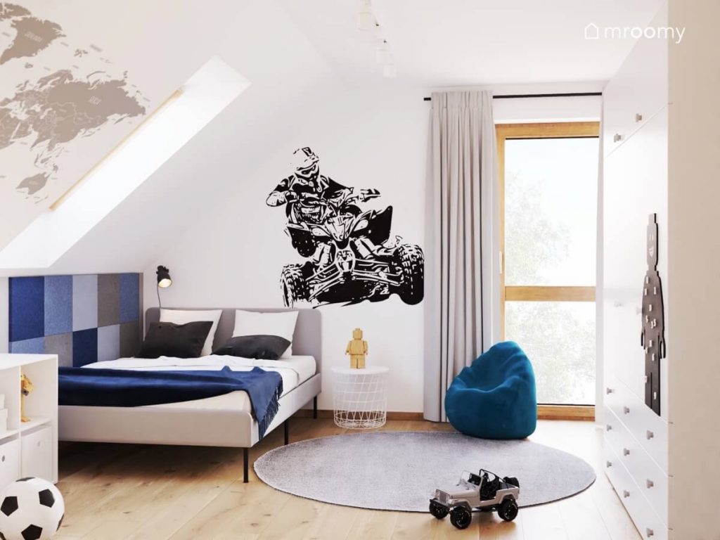 Jasny poddaszowy pokój dla chłopca w wieku szkolnym a w nim szare łóżko uzupełnione kwadratowymi panelami ściennymi niebieska pufa szary dywan i naklejka ścienna z quadem