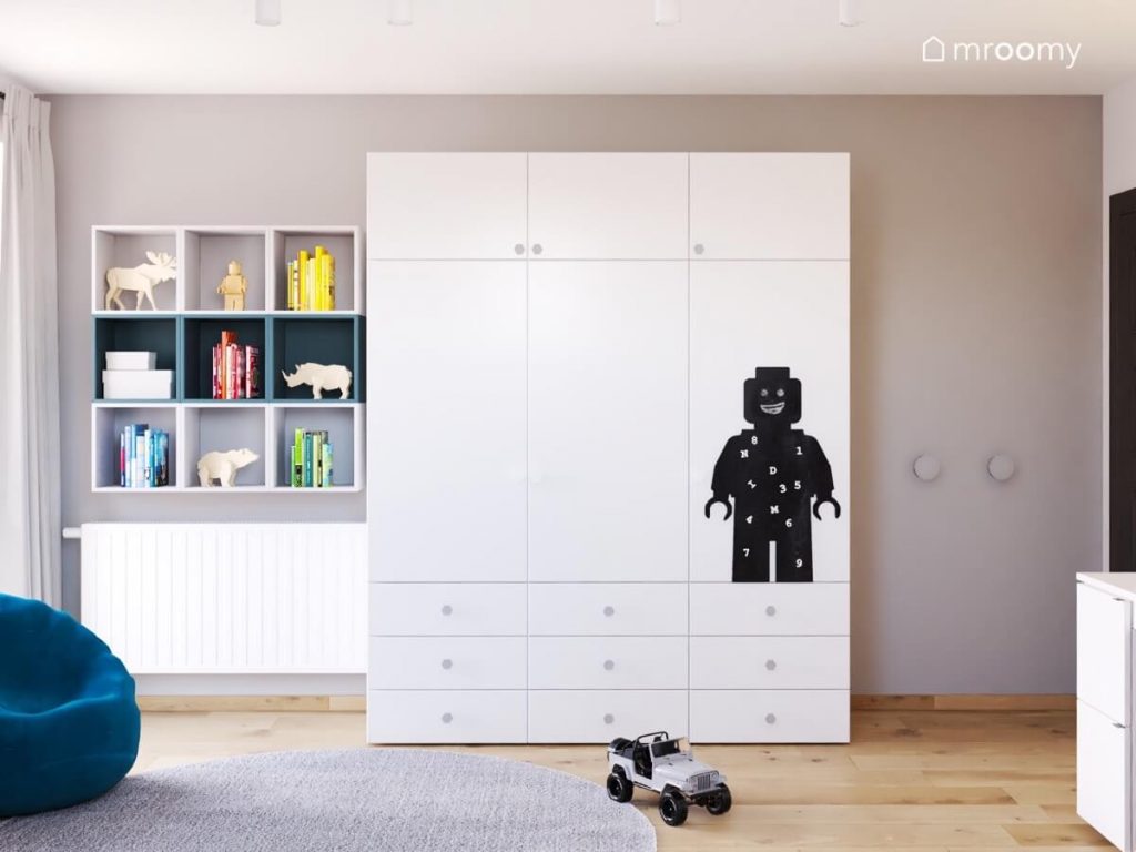 Prosta biała szafa z czarną tablicą kredową w kształcie ludzika Lego a obok dziewięć szafek ściennych w pokoju dla chłopca