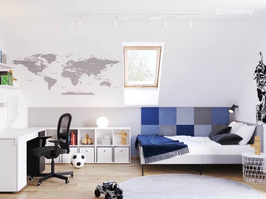 Biało szary poddaszowy pokój dla chłopca w wieku szkolnym a w nim szare łóżko uzupełnione kwadratowymi panelami ściennymi w różnych odcieniach niebieskiego niski regał oraz mapa świata na skosie