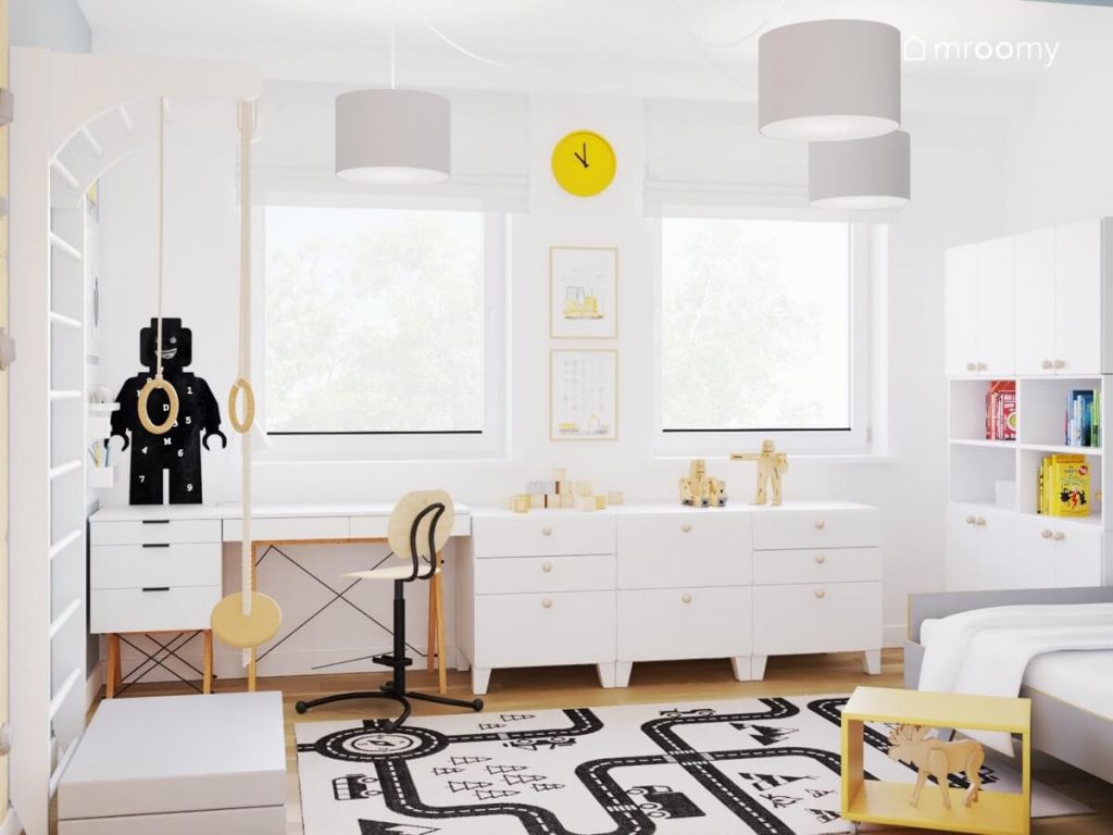 Biała część w biało niebieskim pokoju dla chłopca a w niej białe meble z drewnianymi elementami żółty zegar tablica kredowa w kształcie ludzika Lego a na podłodze dywan z drogą