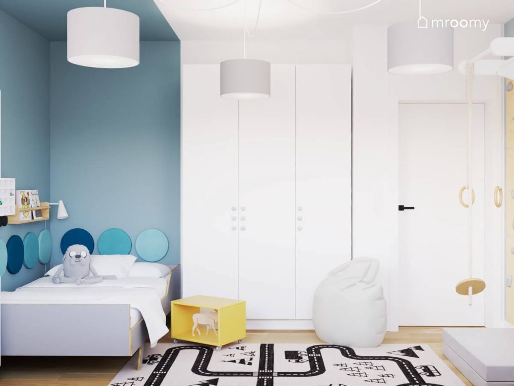 Niebiesko biały pokój dla chłopca a w nim łóżko w części niebieskiej i szafy w części białej a na podłodze żółty stolik nocny na kółkach pufa oraz dywan z drogą
