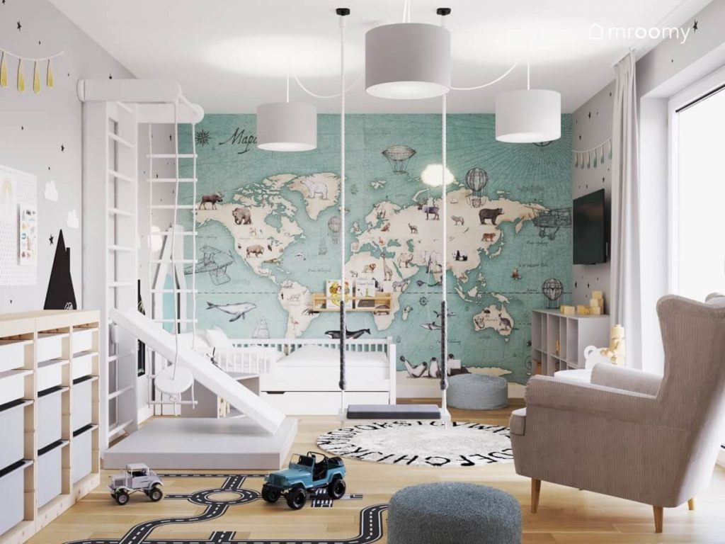 Biało szaro niebieski pokój dla chłopca a w nim białe łóżko biała drabinka gimnastyczna ze zjeżdżalnią huśtawka wisząca szary fotel a na ścianie tapeta z mapą świata