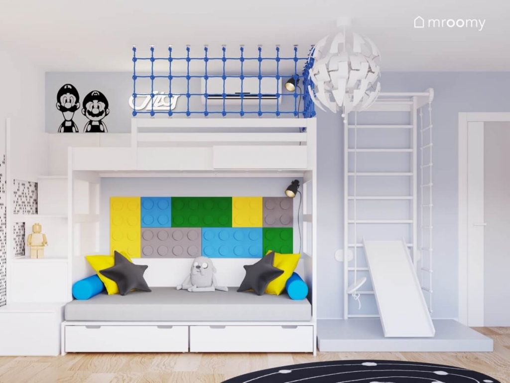 Biała antresola z miejscem do zabawy na górze i łóżkiem uzupełnionym kolorowymi panelami w kształcie klocków na dole a obok biała drabinka gimnastyczna ze zjeżdżalnią w pokoju dla chłopca