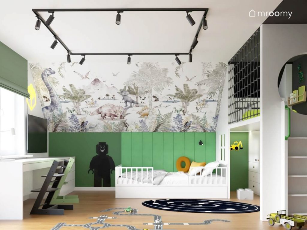 Skos ozdobiony tapetą w dinozaury a pod nim zielona ścianka kolankowa białe łóżko i tablica kredowa w kształcie ludzika Lego a na suficie czarne reflektory w pokoju dla sześciolatka