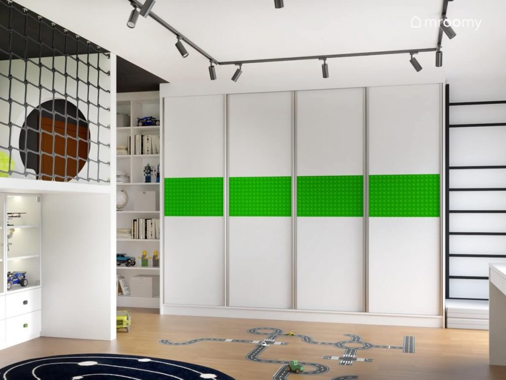 Duża biała szafa z zieloną naklejką ze wzorem klocka Lego a obok czarna drabinka gimnastyczna w pokoju chłopca