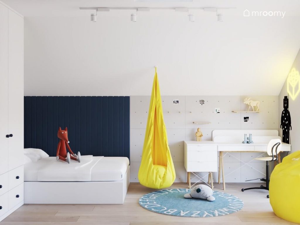 Białe łóżko uzupełnione granatowymi panelami ściennymi białe biurko z kontenerkiem na drewnianych nogach żółty fotel kokon oraz błękitny dywan z alfabetem w pokoju dla chłopca