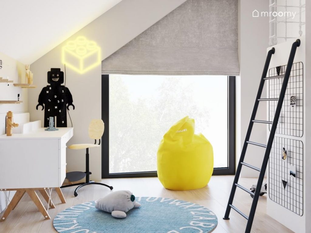 Jasny pokój dla chłopca a w nim białe biurko z krzesłem z podnóżkiem tablica kredowa w kształcie ludzika Lego ledon w kształcie klocka błękitny dywan z alfabetem oraz żółta pufa