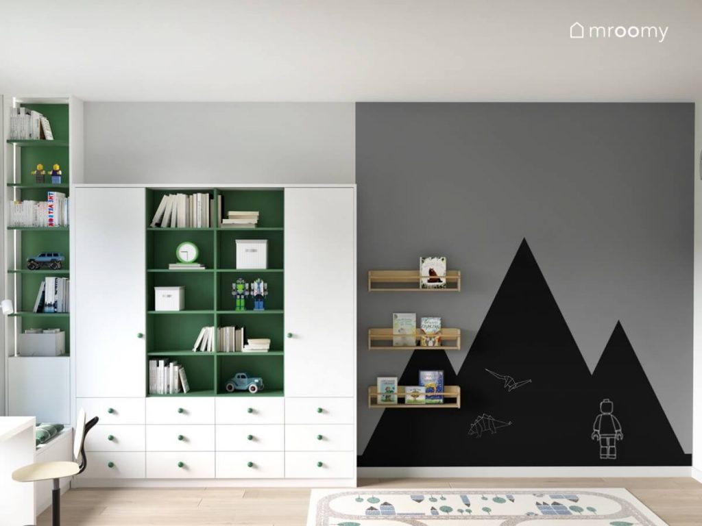 Biało zielona zabudowa meblowa a obok powierzchnia kredowa w kształcie gór i drewniane półki ścienne w pokoju chłopca