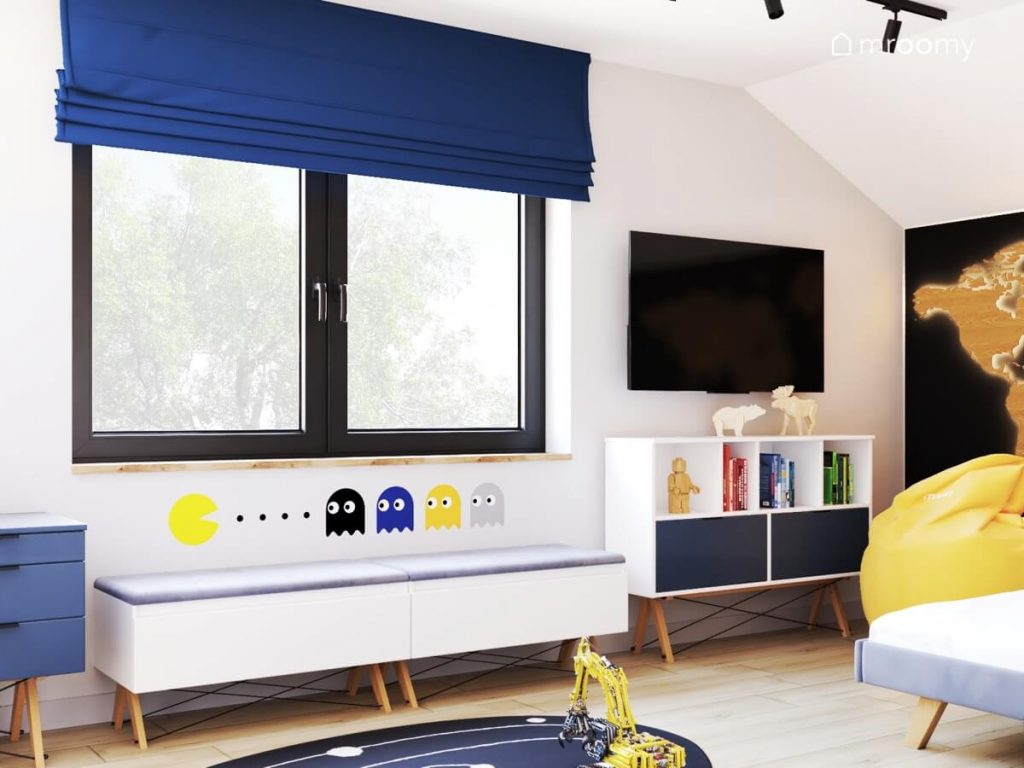 Biało szary poddaszowy pokój dla chłopca a w nim biało granatowe meble telewizor i naklejka z Pacmanem