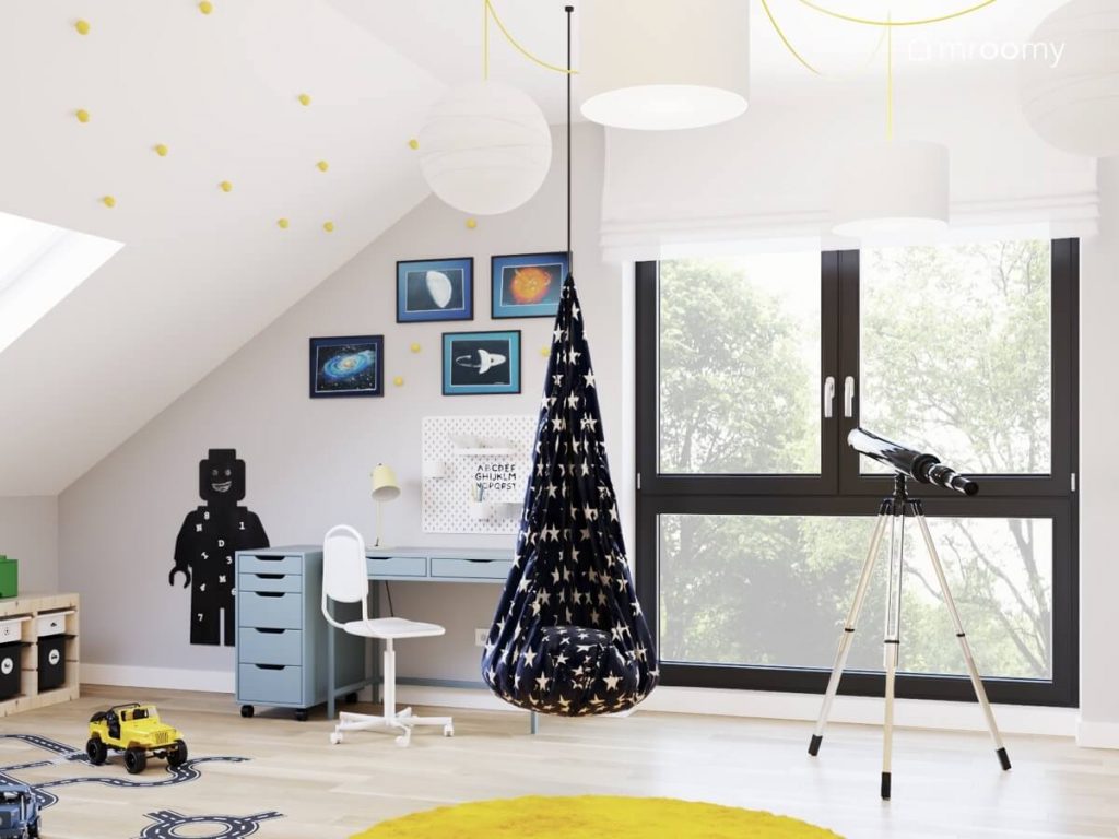 Jasny poddaszowy pokój dla chłopca a w nim teleskop błękitne biurko z kontenerkiem kosmiczne obrazki i tablica kredowa w kształcie ludzika Lego na ścianie a na środku fotel kokon