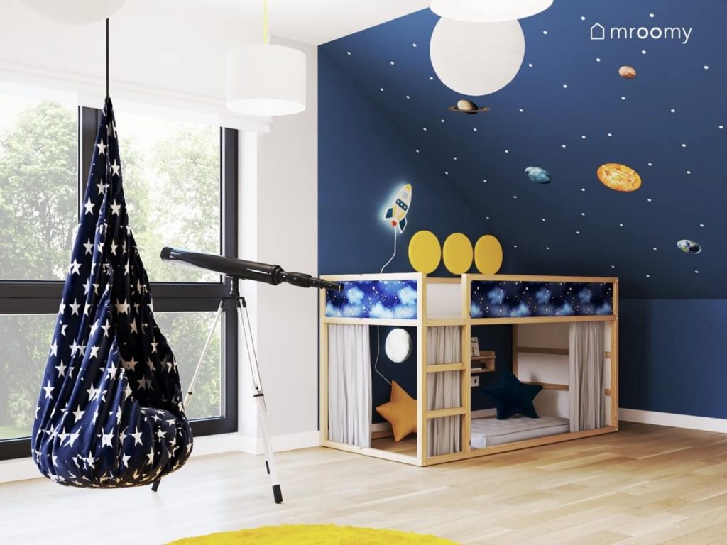 Biało niebieski kosmiczny pokój dla chłopca w wieku przedszkolnym a w nim wiszący fotel kokon teleskop oraz drewniane łóżko