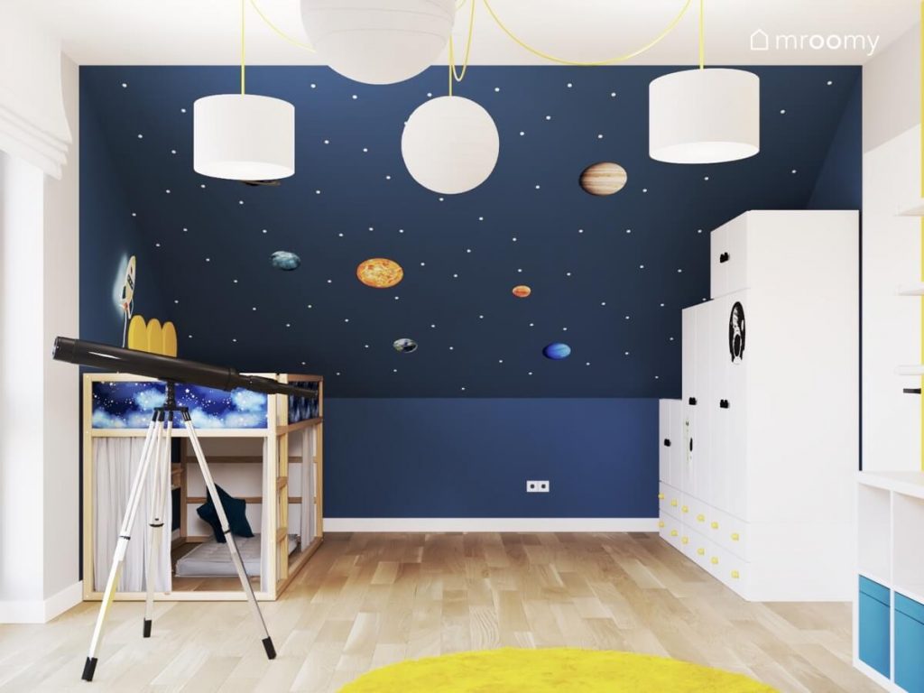 Niebieski skos w białe kropki oklejony planetami a także drewniane łóżko biała szafa teleskop a na suficie białe lampy w pokoju chłopca