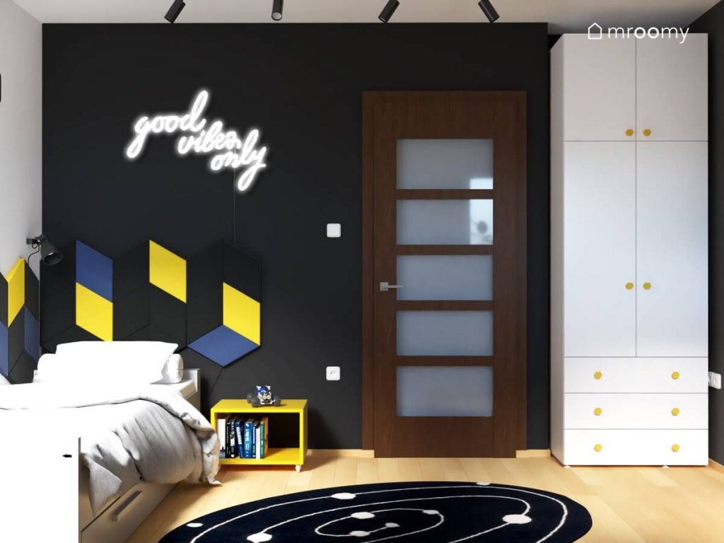 Czarna ściana w pokoju dla chłopca w wieku szkolnym z na niej ledon w kształcie napisu kompozycja z paneli ściennych oraz biała szafa z żółtymi gałkami