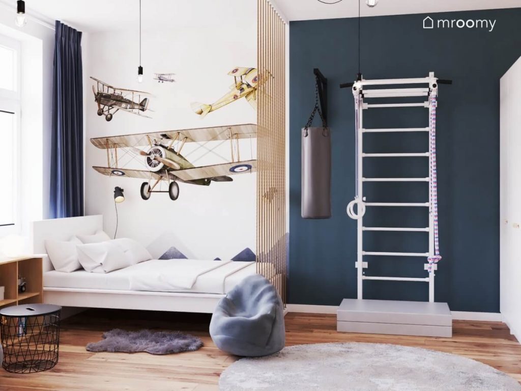 Białe łóżko a na ścianie nad nim samoloty a obok worek treningowy i drabinka gimnastyczna z materacem w pokoju nastolatka